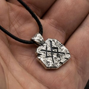 May include: A silver pendant necklace with a textured, geometric design. The pendant features a stylized symbol with a rough, stone-like appearance.