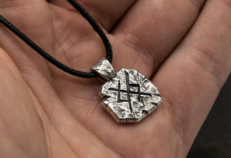 May include: A silver pendant necklace with a textured, geometric design. The pendant features a stylized symbol with a rough, stone-like appearance.