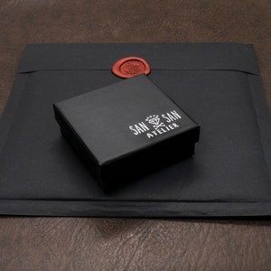May include: A black square box with the text "SAN SAN ATELIER" in white lettering. The box is sitting on a black envelope with a red wax seal.