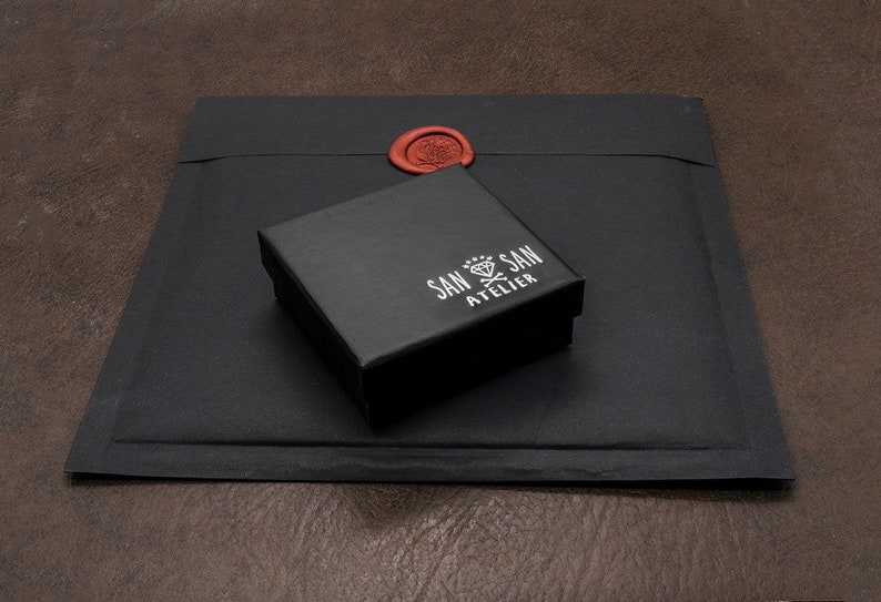 May include: A black square box with the text "SAN SAN ATELIER" in white lettering. The box is sitting on a black envelope with a red wax seal.