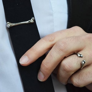May include: A silver tie clip and ring in the shape of a bone. The tie clip is worn on a black tie and the ring is worn on a finger.
