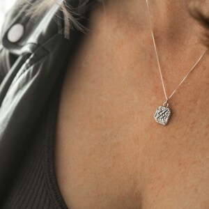 May include: A silver necklace with a square pendant featuring a textured design. The pendant is hanging on a delicate chain.