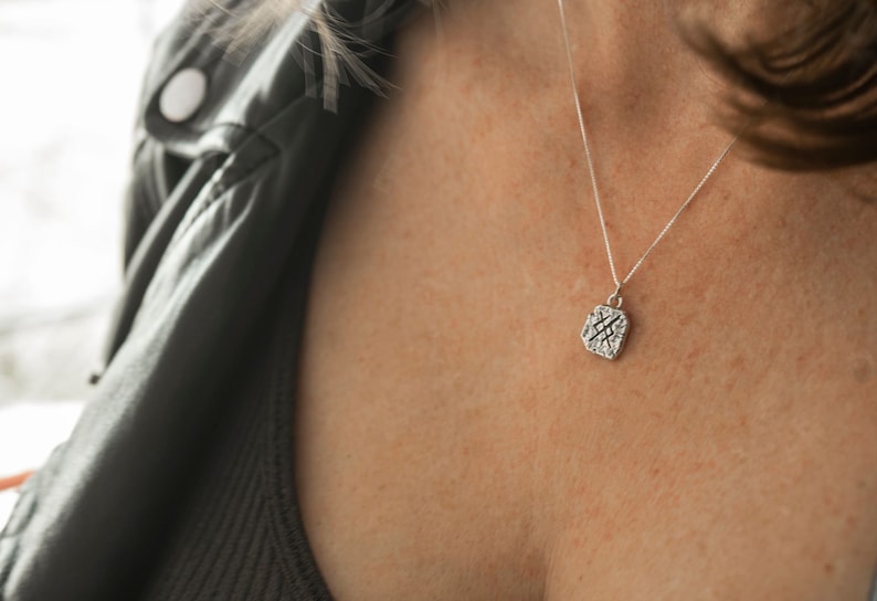 May include: A silver necklace with a square pendant featuring a textured design. The pendant is hanging on a delicate chain.