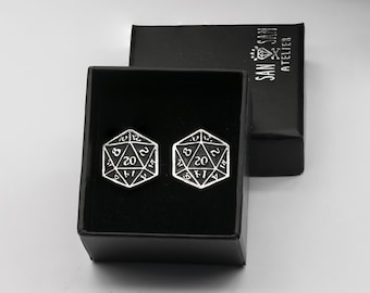 May include: A pair of silver-colored cufflinks in the shape of twenty-sided dice, each displaying the number 20. The cufflinks are presented in a black box with the text "SAN SAN ATELIER" printed on the lid.