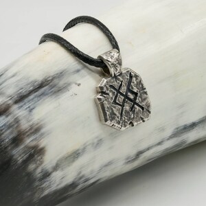 May include: A silver pendant necklace with a textured, geometric design and a black leather cord. The pendant features a stylized symbol.