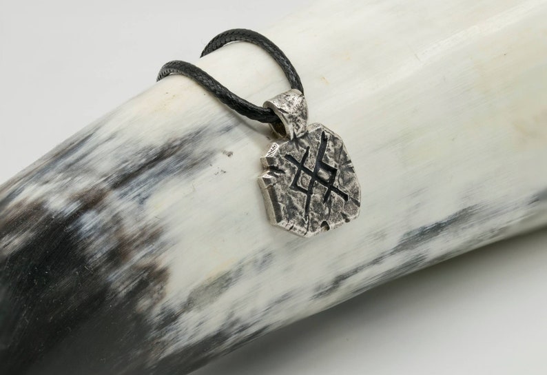 May include: A silver pendant necklace with a textured, geometric design and a black leather cord. The pendant features a stylized symbol.