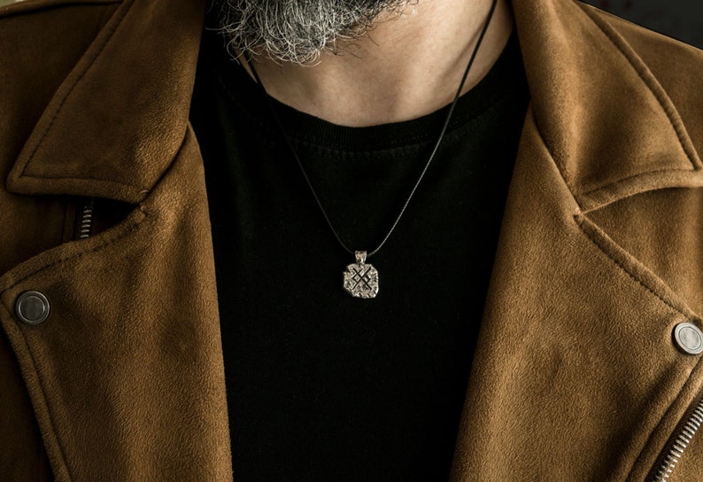 May include: A silver pendant necklace with a detailed design hangs from a black cord. The necklace is worn by a person wearing a brown suede jacket and a black t-shirt.