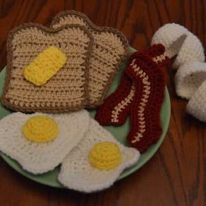 May include: Crocheted breakfast food set with two slices of toast, two eggs, and two strips of bacon on a green plate.