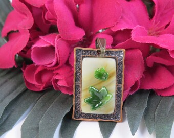 Può includere: Un ciondolo rettangolare con un centro in vetro color crema e due accenti lucidi a forma di foglia verde. Il pezzo è racchiuso in una cornice di metallo scuro, testurizzata e decorata, con un gancio metallico abbinato sulla parte superiore.