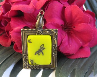 Può includere: Ciondolo quadrato con cornice color ottone e centro giallo brillante. Il centro presenta un disegno di libellula nera. Il ciondolo è sospeso da un'ansa color ottone. Lo sfondo comprende un vibrante fiore rosa e foglie verdi.
