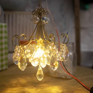 May include: Miniature chandelier with a gold-colored frame and clear crystal accents. The chandelier is lit, casting a warm glow. The crystals are teardrop and round shaped. The fixture is suspended by a chain. The background is blurred.