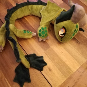 May include: Green plush dragon toy with a long tail and wings. The dragon has a green and black body with a grey head and orange eyes.