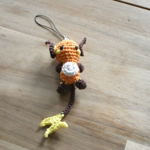 Puede incluir: Un llavero Eevee de crochet naranja y marrón con vientre blanco y cola amarilla. El llavero tiene un cordón negro.