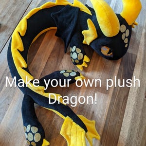 May include: A black and yellow plush dragon with the text "Make your own plush Dragon!" and "DIY sewing Kit". The dragon features yellow horns, claws, and accents. The dragon is displayed on a wooden surface.