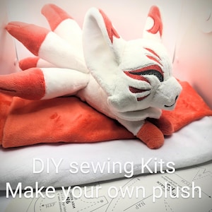 May include: A plush, white and orange fox with multiple tails, lying on a white and orange surface. The fox has red markings around its eyes and on its ears. The image includes the text "DIY sewing Kits Make your own plush".