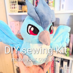 May include: A light blue plush bird with a gray beak and red accents around the eyes. The image includes the text "DIY sewing Kit make your own plush". The bird has large wings and a fluffy appearance.