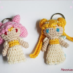 May include: Two crocheted doll keychains, one with pink hair and a yellow dress, the other with yellow hair and a yellow dress. Both dolls have blue eyes and are wearing white dresses.