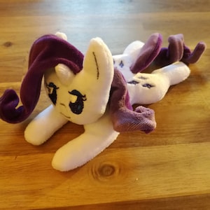May include: A white plush toy pony with a purple mane and tail. The pony has blue eyes and a white horn. The pony is lying on its side with its head resting on its front leg.
