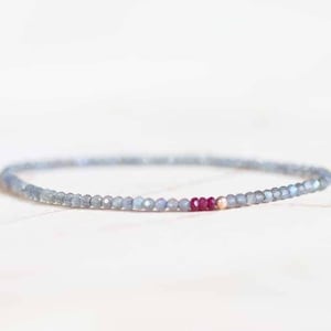 May include: A delicate bracelet featuring small, faceted, light gray beads with a few vibrant red beads. The bracelet is strung on a thin, elastic cord, creating a minimalist and elegant design.