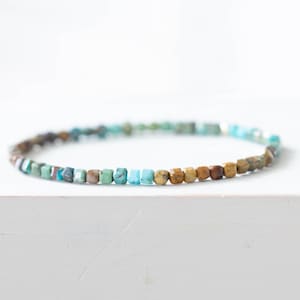 Könnte beinhalten: Ein zartes Armband mit kleinen, quadratisch geschnittenen Perlen in Türkis, Braun und Beige. Die Perlen sind zu einem flexiblen Band aufgereiht, ideal für den täglichen Gebrauch. Das Armband wird auf einer weißen Oberfläche präsentiert.