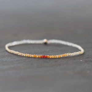 May include: A delicate beaded bracelet with small, round beads in clear, yellow, orange, and red. The bracelet is strung on a thin cord and designed to be worn around the wrist. The bracelet's total length is approximately 17.8 cm.