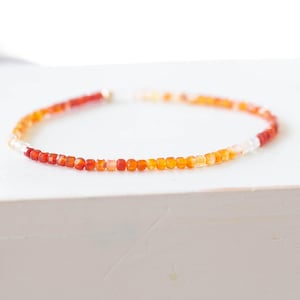 May include: A delicate bracelet featuring small, square beads in shades of red, orange, and white. The beads are strung together to form a circular shape. The bracelet is displayed on a white surface.