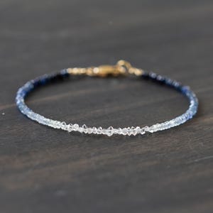 May include: A delicate bracelet featuring a gradient of blue and clear gemstones. The bracelet has a gold clasp and is displayed on a dark wooden surface. The gemstones transition from dark blue to clear.