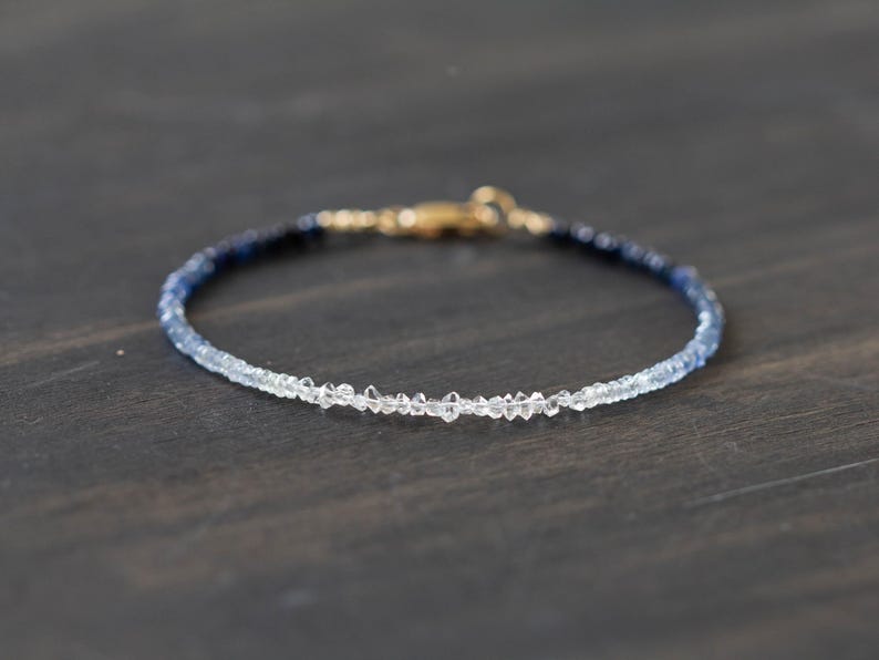 May include: A delicate bracelet featuring a gradient of blue and clear gemstones. The bracelet has a gold clasp and is displayed on a dark wooden surface. The gemstones transition from dark blue to clear.