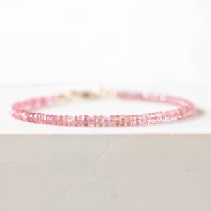 Op de afbeelding: Een delicate roze edelsteenarmband. De armband heeft kleine, ronde, roze edelstenen die aan elkaar zijn geregen met een goudkleurige sluiting. De armband wordt gepresenteerd op een wit oppervlak, met zacht, diffuus licht.