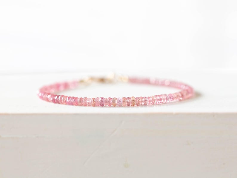 Op de afbeelding: Een delicate roze edelsteenarmband. De armband heeft kleine, ronde, roze edelstenen die aan elkaar zijn geregen met een goudkleurige sluiting. De armband wordt gepresenteerd op een wit oppervlak, met zacht, diffuus licht.