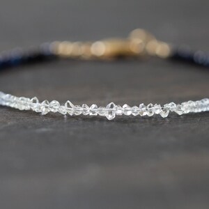 May include: A delicate bracelet featuring a gradient of blue and clear gemstones. The bracelet has a gold clasp and is set against a dark background. The gemstones appear to be small, faceted crystals.