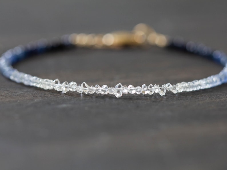May include: A delicate bracelet featuring a gradient of blue and clear gemstones. The bracelet has a gold clasp and is set against a dark background. The gemstones appear to be small, faceted crystals.