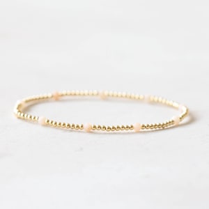 Pode incluir: Uma delicada pulseira de contas douradas com pequenas contas redondas douradas e intercaladas com contas maiores de cor pêssego claro. A pulseira é exibida em uma superfície branca.