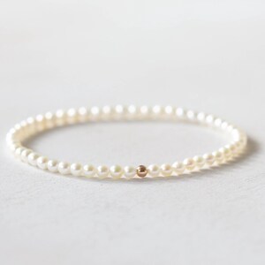 May include: A delicate bracelet composed of small, round, creamy white pearls. A single, small gold-colored bead is centered on the bracelet. The bracelet is set against a soft, white background.