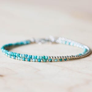 Puede incluir: Una delicada pulsera con dos hilos de pequeñas cuentas de color turquesa y cuentas plateadas. La pulsera se exhibe sobre una superficie de madera clara, mostrando su elegancia sencilla y su artesanía.