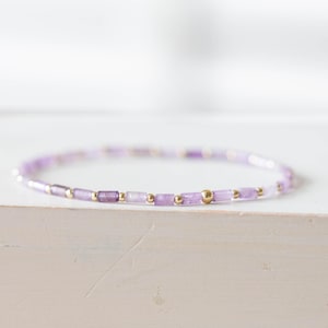 May include: A delicate bracelet featuring small, rectangular lavender-colored beads interspersed with gold-colored beads. The bracelet is displayed on a white surface, showcasing its minimalist design and color palette.