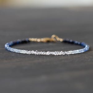 May include: Delicate bracelet featuring a gradient of blue and clear beads. The beads transition from dark blue to clear, connected by a gold clasp. The bracelet is a fine piece of jewelry.