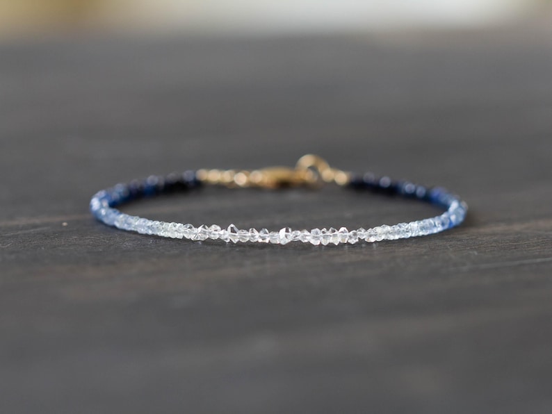 May include: Delicate bracelet featuring a gradient of blue and clear beads. The beads transition from dark blue to clear, connected by a gold clasp. The bracelet is a fine piece of jewelry.