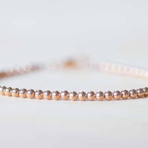 May include: A delicate bracelet featuring small, evenly spaced, rose gold beads. The bracelet is strung on a thin, flexible band, creating a minimalist and elegant design. The beads have a polished, reflective surface.