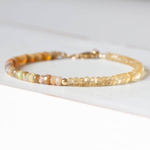 Puede incluir: Una pulsera delicada que presenta una hilera de cuentas de ópalo facetadas y multicolores en un lado y cuentas de citrino translúcidas de color amarillo pálido en el otro, separadas por una pequeña cuenta dorada y un cierre de mosquetón.