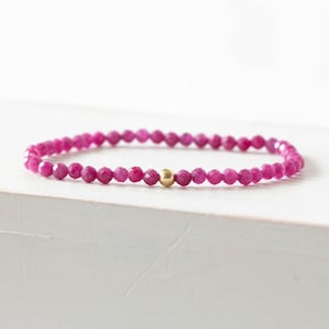 May include: A beaded bracelet featuring small, faceted, deep pink gemstone beads arranged in a continuous circular pattern, accented by a single, smooth, polished gold-toned spherical bead positioned at the center of the strand.