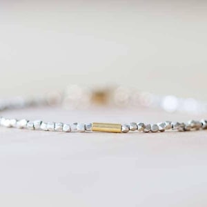 Pode incluir: Uma pulseira delicada com pequenas contas prateadas e um detalhe cilíndrico dourado. A pulseira é enfiada em um cordão fino e flexível e é exibida em uma superfície clara.