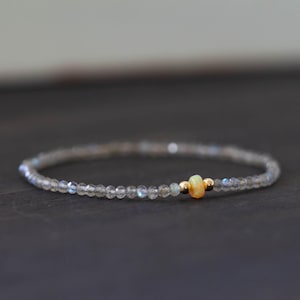 Peut inclure: Un bracelet délicat composé de petites perles claires et facettées. Le bracelet est orné d'une petite pierre précieuse jaune-orangé et de deux perles dorées. Le bracelet est présenté sur une surface sombre.