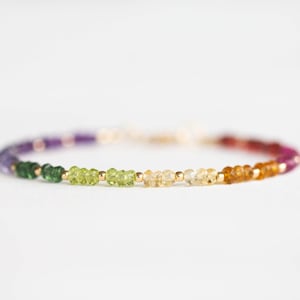 May include: A beaded bracelet featuring a spectrum of colors, including purple, green, yellow, orange, and red. The bracelet is made with small, round gemstones and gold-colored beads. The gemstones are arranged in a gradient pattern.