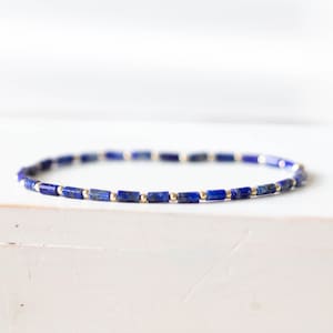 Puede incluir: Una pulsera delicada con pequeñas cuentas rectangulares de lapislázuli azul oscuro intercaladas con cuentas doradas. La pulsera se exhibe sobre una superficie blanca, mostrando su diseño minimalista y su elegante contraste de colores.