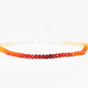 May include: A delicate beaded bracelet featuring a gradient of colors, transitioning from white to yellow, orange, and deep red. The small, faceted beads are strung together, creating a subtle and elegant accessory.