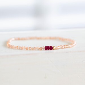 May include: A delicate bracelet featuring small, peach-colored beads. The bracelet is accented with three deep red beads and a small gold bead. The jewelry is displayed on a white surface.