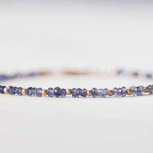 May include: Delicate bracelet featuring small, faceted, blue-violet gemstones interspersed with small, round, gold-colored beads. The bracelet is strung on a thin, flexible cord, creating a minimalist and elegant design.