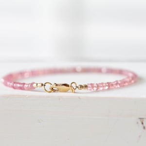 Op de afbeelding: Een delicate roze edelsteenarmband met een goudkleurige sluiting. De armband is gemaakt van kleine, gelijkmatig gevormde roze kralen. De sluiting is een eenvoudig haak- en oogontwerp. De armband wordt op een wit oppervlak weergegeven.