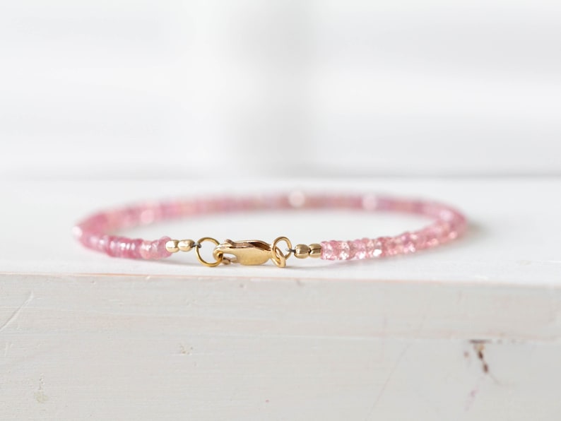 Op de afbeelding: Een delicate roze edelsteenarmband met een goudkleurige sluiting. De armband is gemaakt van kleine, gelijkmatig gevormde roze kralen. De sluiting is een eenvoudig haak- en oogontwerp. De armband wordt op een wit oppervlak weergegeven.